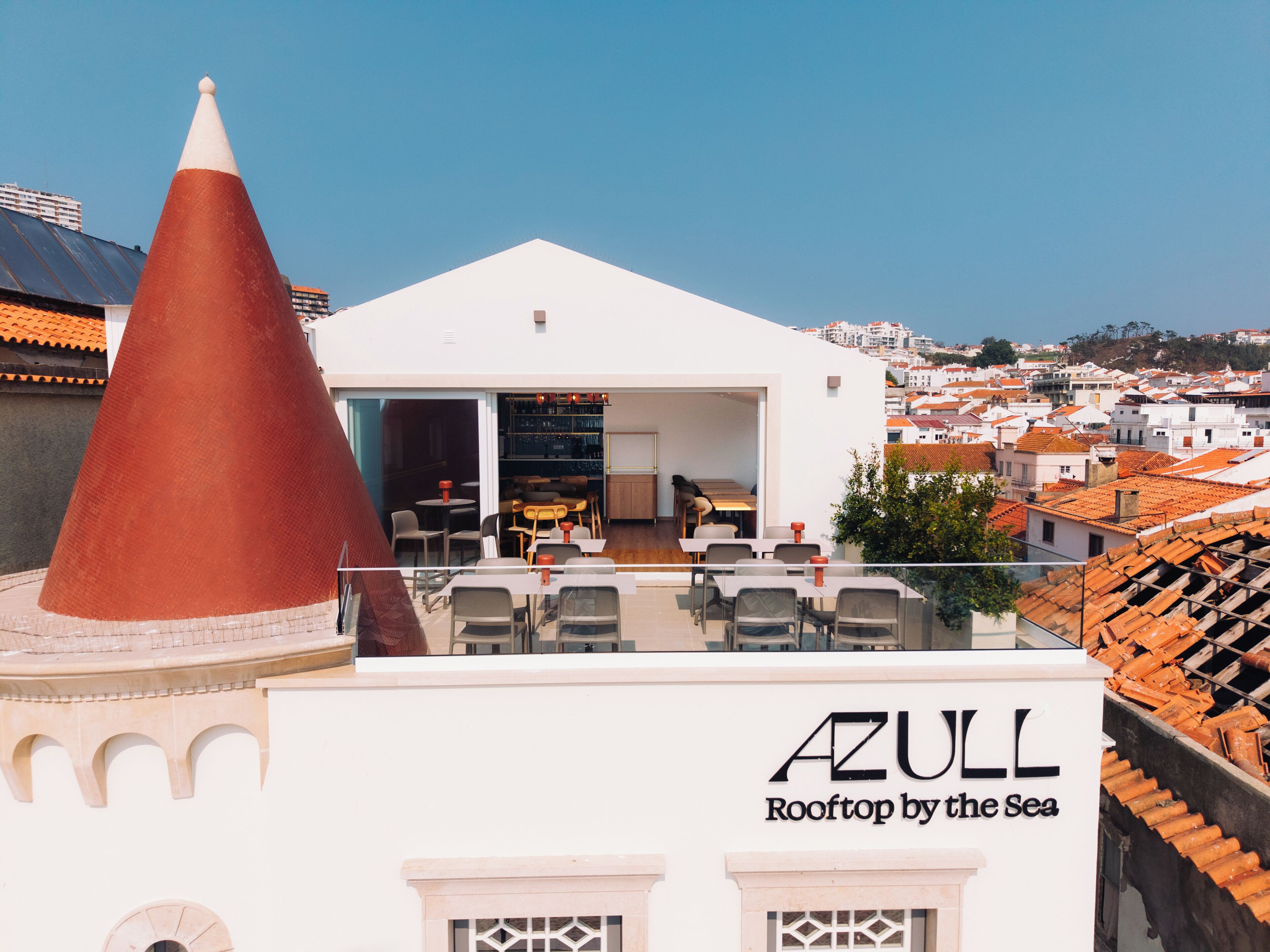 A rooftop terrace with tables and chairs, a large conical structure, and a view of a coastal town.