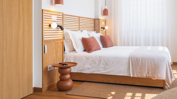 A modern bedroom with a wooden headboard, a bedside table with a remote, and a large window with sheer curtains.