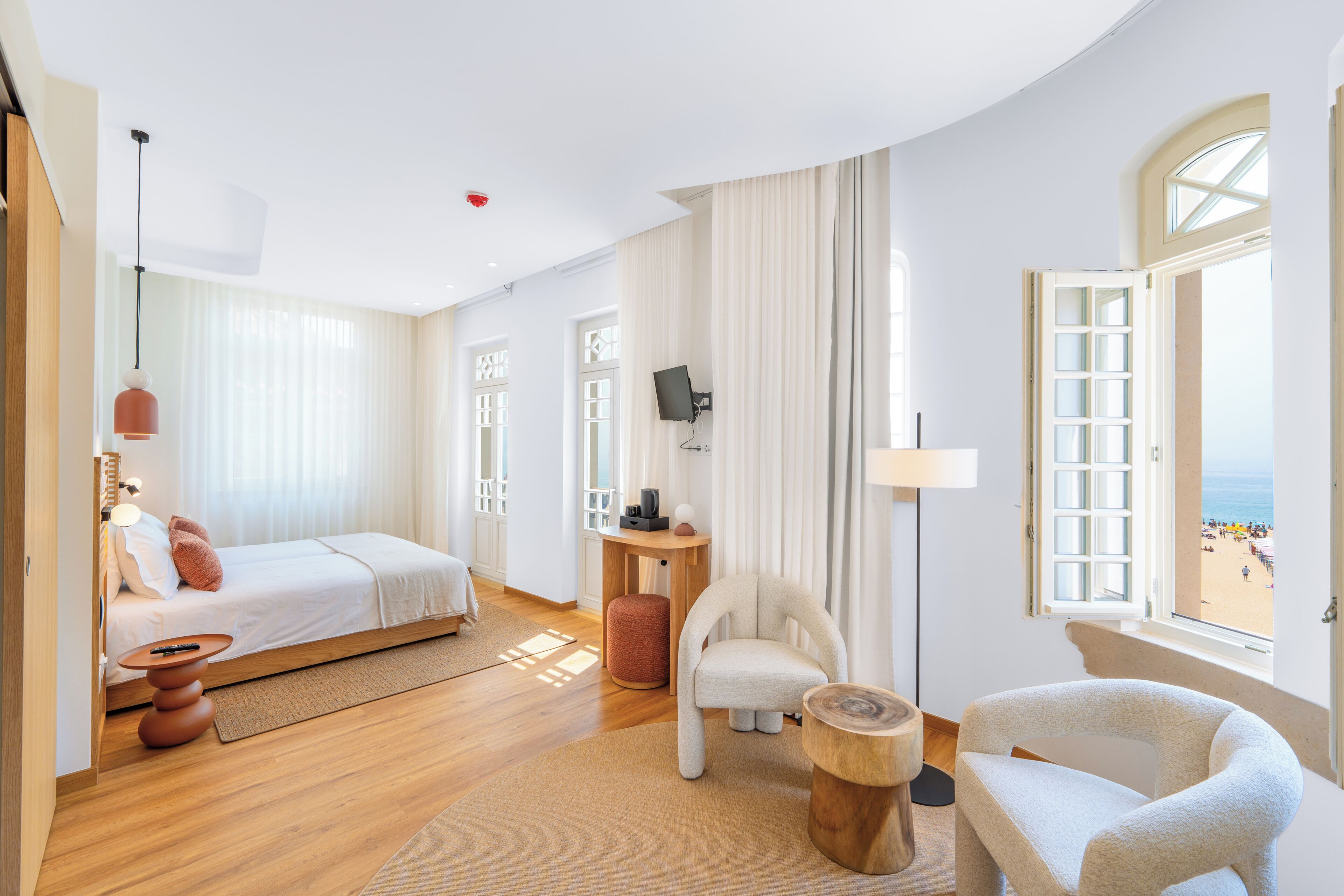 Ein Hotelzimmer mit Bett, Sessel, kleinem Tisch, Fernseher und Blick auf den Strand.