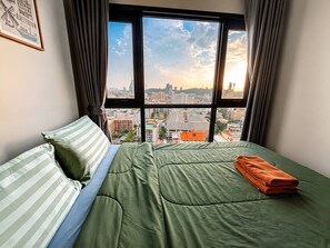 Una cama con ropa de cama verde y almohadas a rayas, una ventana con vista a la ciudad y un cuadro en la pared.