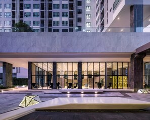 Une entrée d’hôtel moderne avec une façade en verre, un espace extérieur aménagé pour s’asseoir et une vue sur les bâtiments environnants.