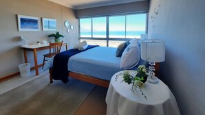A bedroom with a bed, bedside table, and a desk with a chair. There are windows with a view of the beach. - Amazing View For Less Bloubergstrand Beachfront (Cape Town)