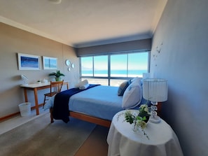 A bedroom with a bed, bedside table, and a desk with a chair. There are windows with a view of the beach. - Amazing View For Less Bloubergstrand Beachfront (Cape Town)