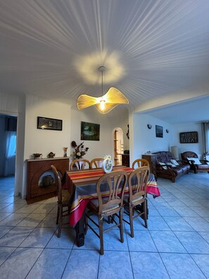 A dining area with a table set for four, a hanging light fixture, and a sofa in the background. - Villa la Hutte, Grand Piquey, Lège, Cap Ferret (Lège-Cap-Ferret)
