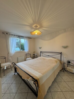 A bedroom with a bed, a chair, a table, and a window with curtains. - Villa la Hutte, Grand Piquey, Lège, Cap Ferret (Lège-Cap-Ferret)