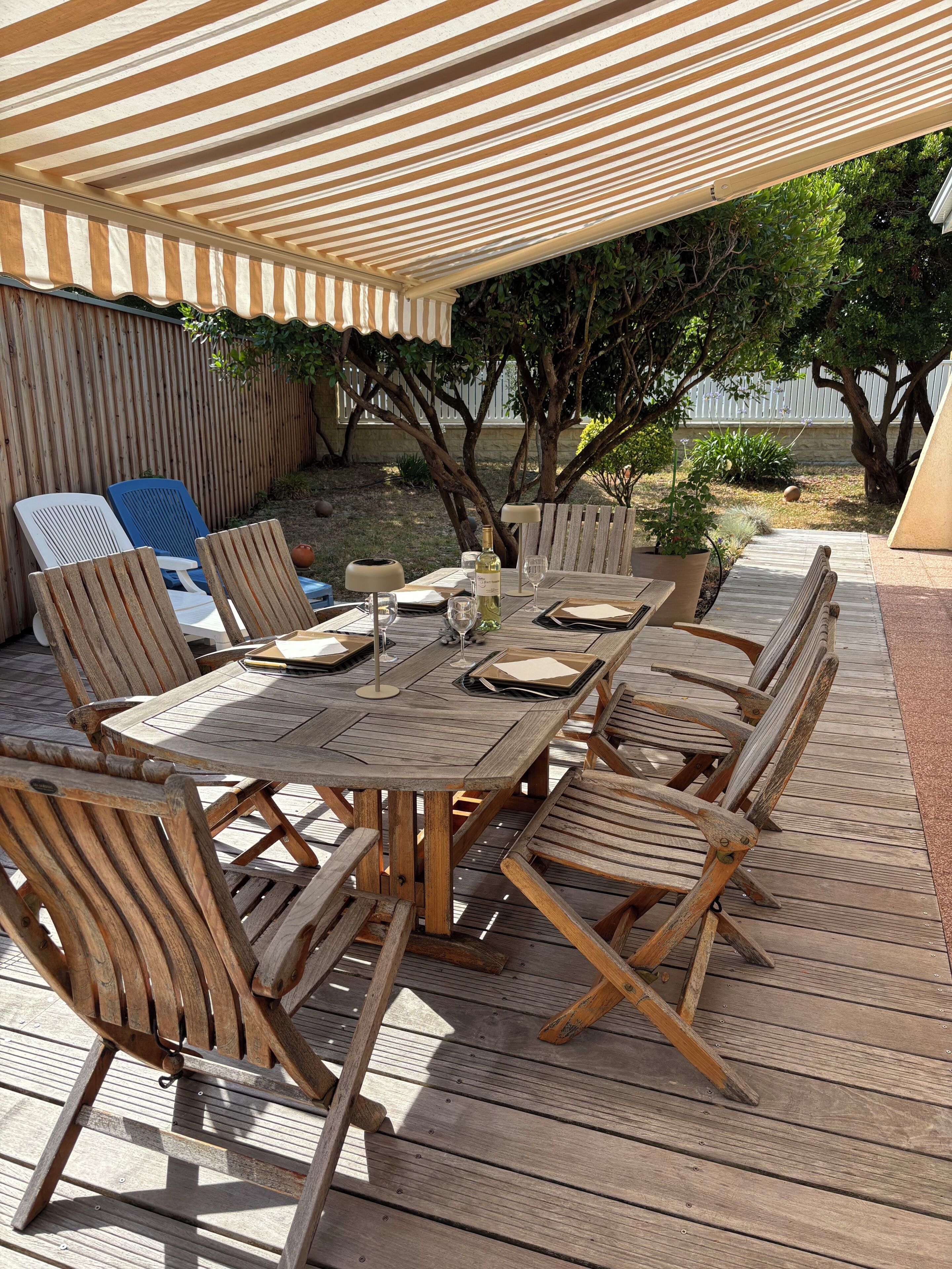 An outdoor patio with a wooden table and chairs, a striped awning overhead, and a view of the surrounding garden.