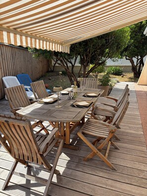 An outdoor patio with a wooden table and chairs, a striped awning overhead, and a view of the surrounding garden. - Villa la Hutte, Grand Piquey, Lège, Cap Ferret (Lège-Cap-Ferret)