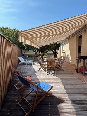 Una terraza exterior con suelo de madera, una zona de estar cubierta con sillas y una mesa, y una cerca de madera que delimita el espacio.