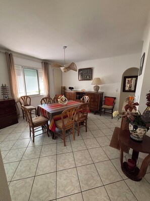 A dining area with a round table, chairs, a ceiling fan, a window with curtains, and a wall-mounted lamp. - Villa la Hutte, Grand Piquey, Lège, Cap Ferret (Lège-Cap-Ferret)