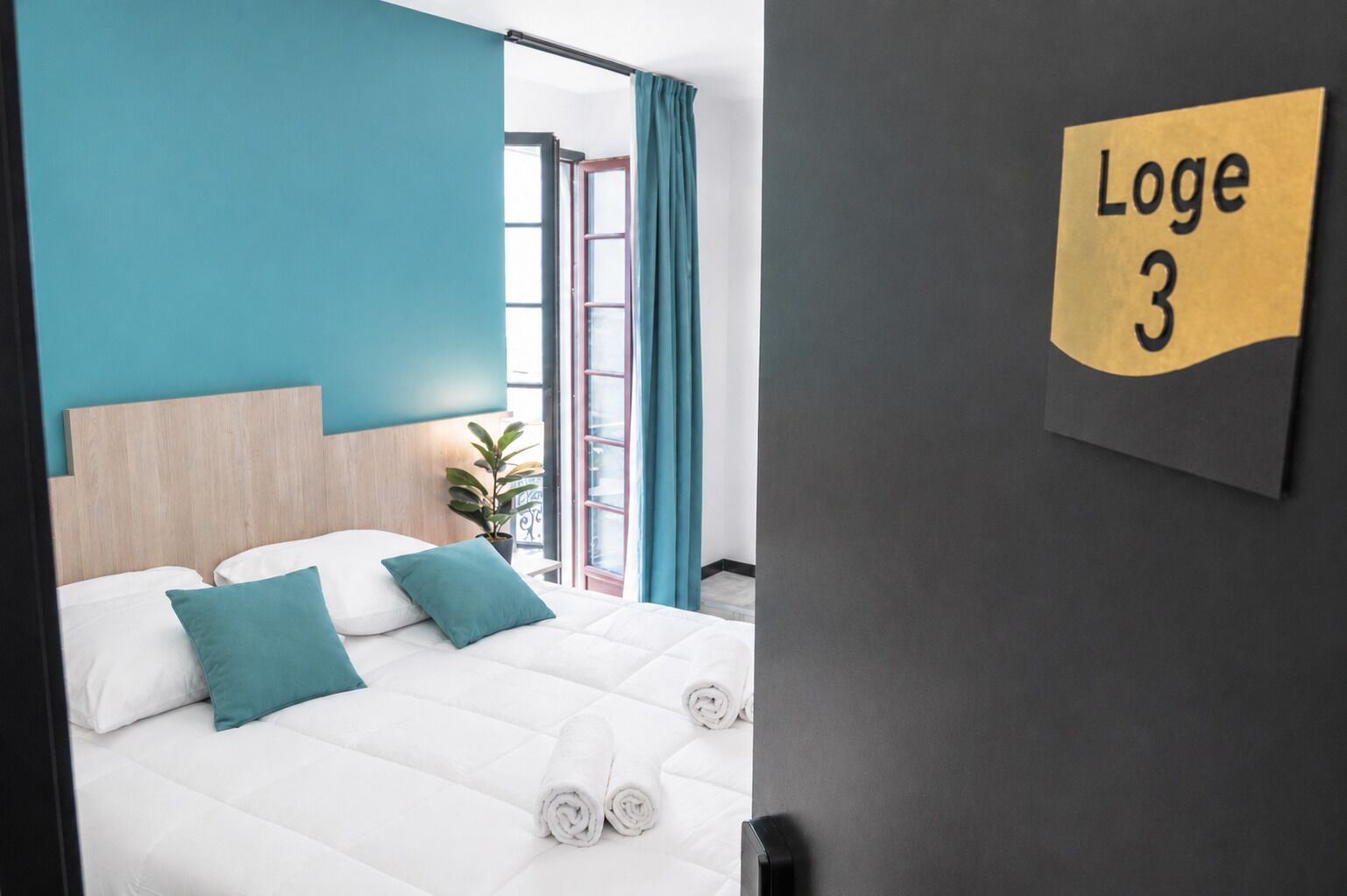A hotel room with a bed, teal pillows, a window with curtains, and a sign indicating 'Loge 3'.