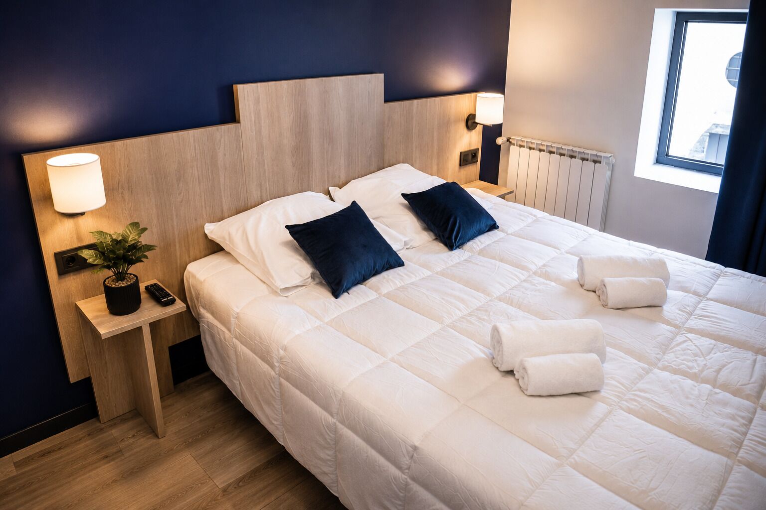 A hotel room with a large bed, two bedside lamps, a small table with a plant and remote, and a window with curtains.