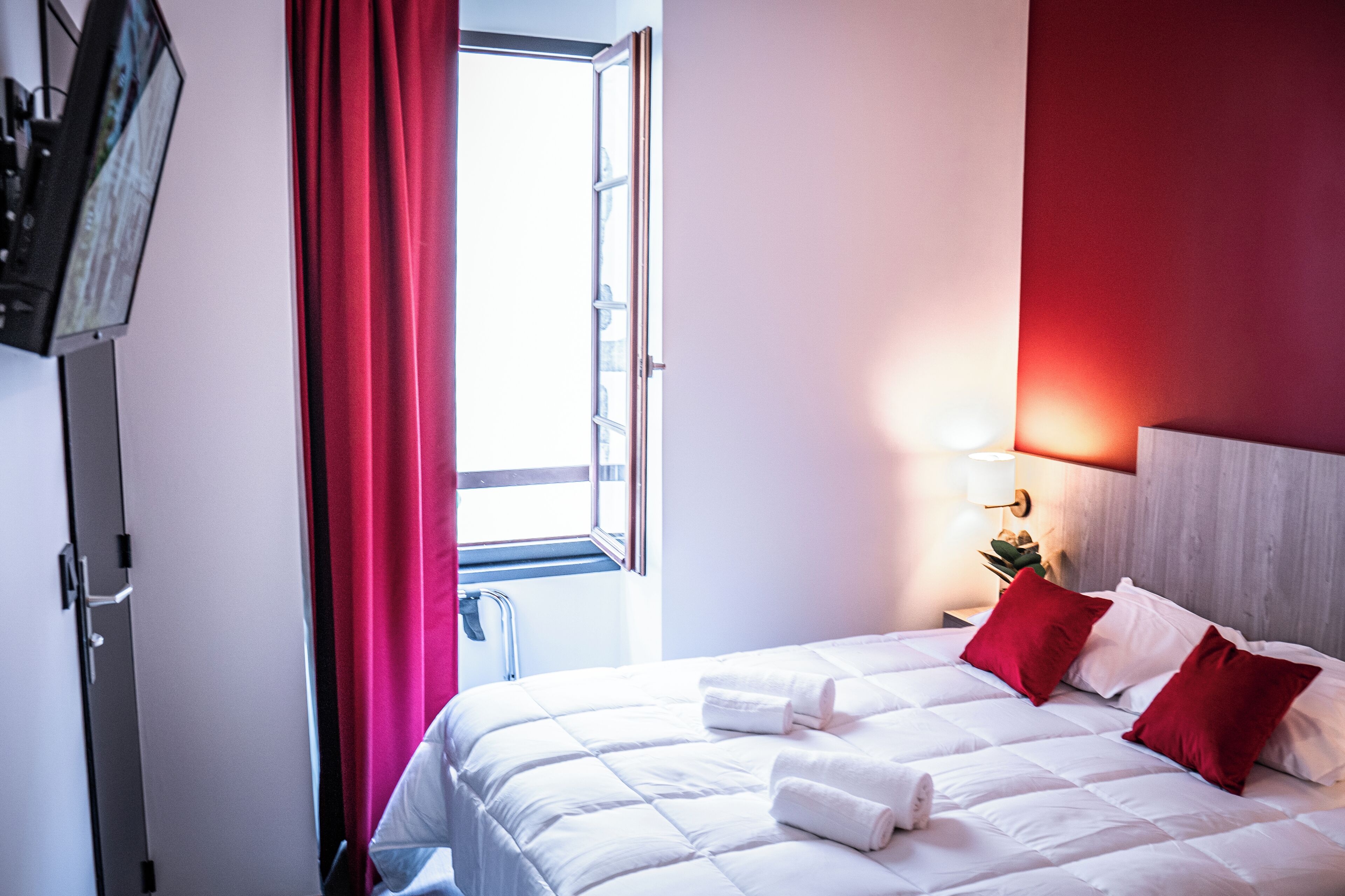 A hotel room with a bed, a TV, a window with curtains, and a nightstand with a lamp.
