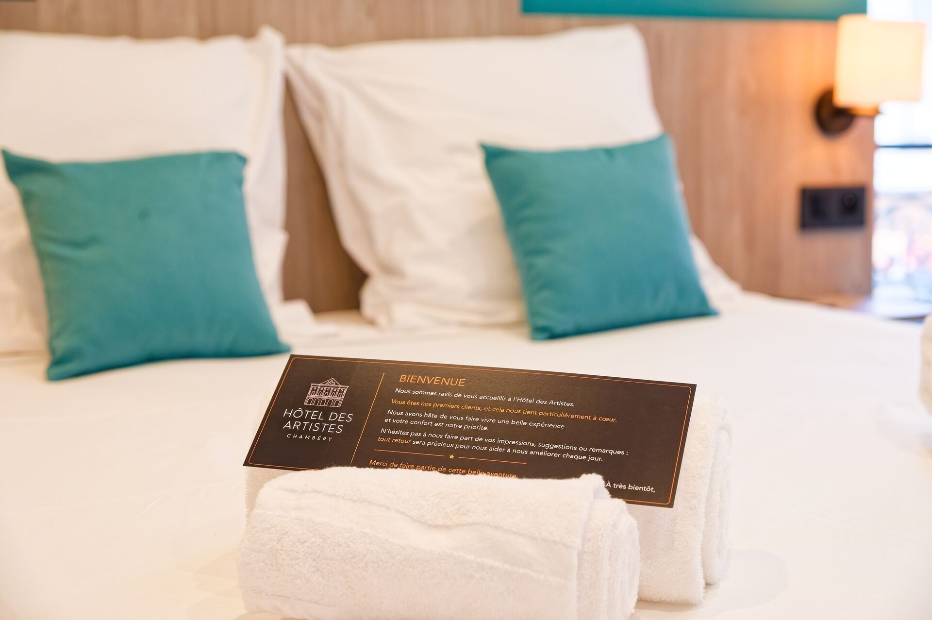A hotel room with a neatly made bed, two teal pillows, and a welcome card on the bed.