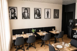 Un restaurante con asientos tapizados en negro, mesas de madera y sillas negras. Se observan cuadros enmarcados en la pared y una máquina de café en una esquina.