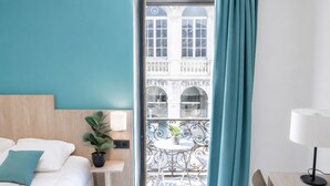Habitación de hotel con cama, una mesita con una planta, balcón con vista a un edificio y una silla.