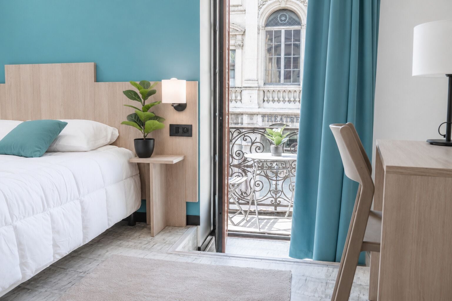 Une chambre d’hôtel avec un lit, une table de chevet avec une lampe, une plante en pot et une vue sur un balcon doté d’une balustrade.