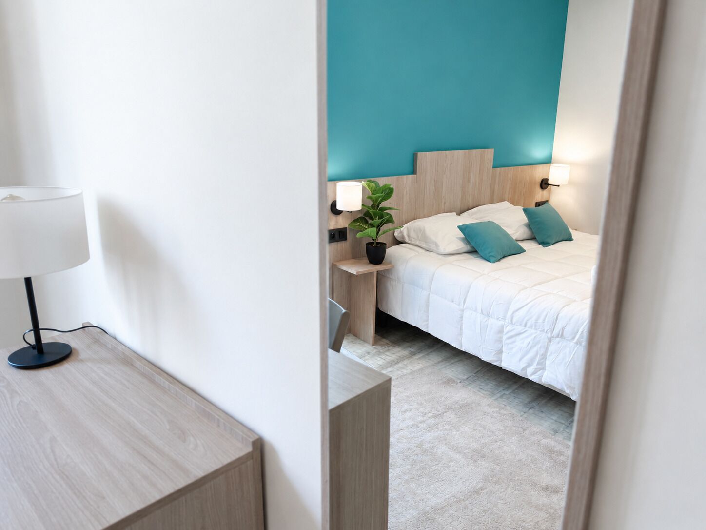 Una habitación de hotel con cama, mesita de noche, lámpara y una planta en maceta.