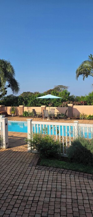 A swimming pool area with a brick patio, lounge chairs, and a table with an umbrella. - Aloe Self Catering One Bedroom With Kitchenette (Port Shepstone)