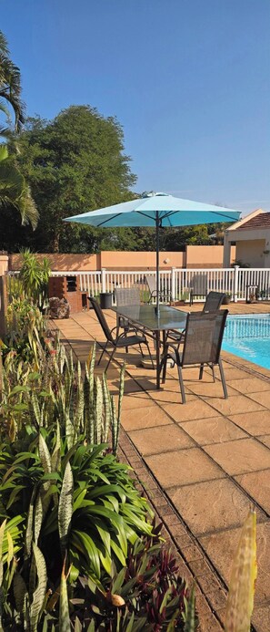 A poolside area with a glass table and chairs, surrounded by a white fence, and a large umbrella providing shade.