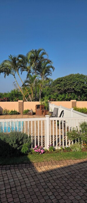 A paved walkway leading to a white fence with a bench and a brick structure, surrounded by palm trees and greenery.