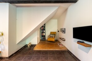Ein modernes Wohnzimmer mit schräg verlaufender Decke, einer Holzbank, einem Bücherregal, einem Fernseher und einem kleinen Hund.