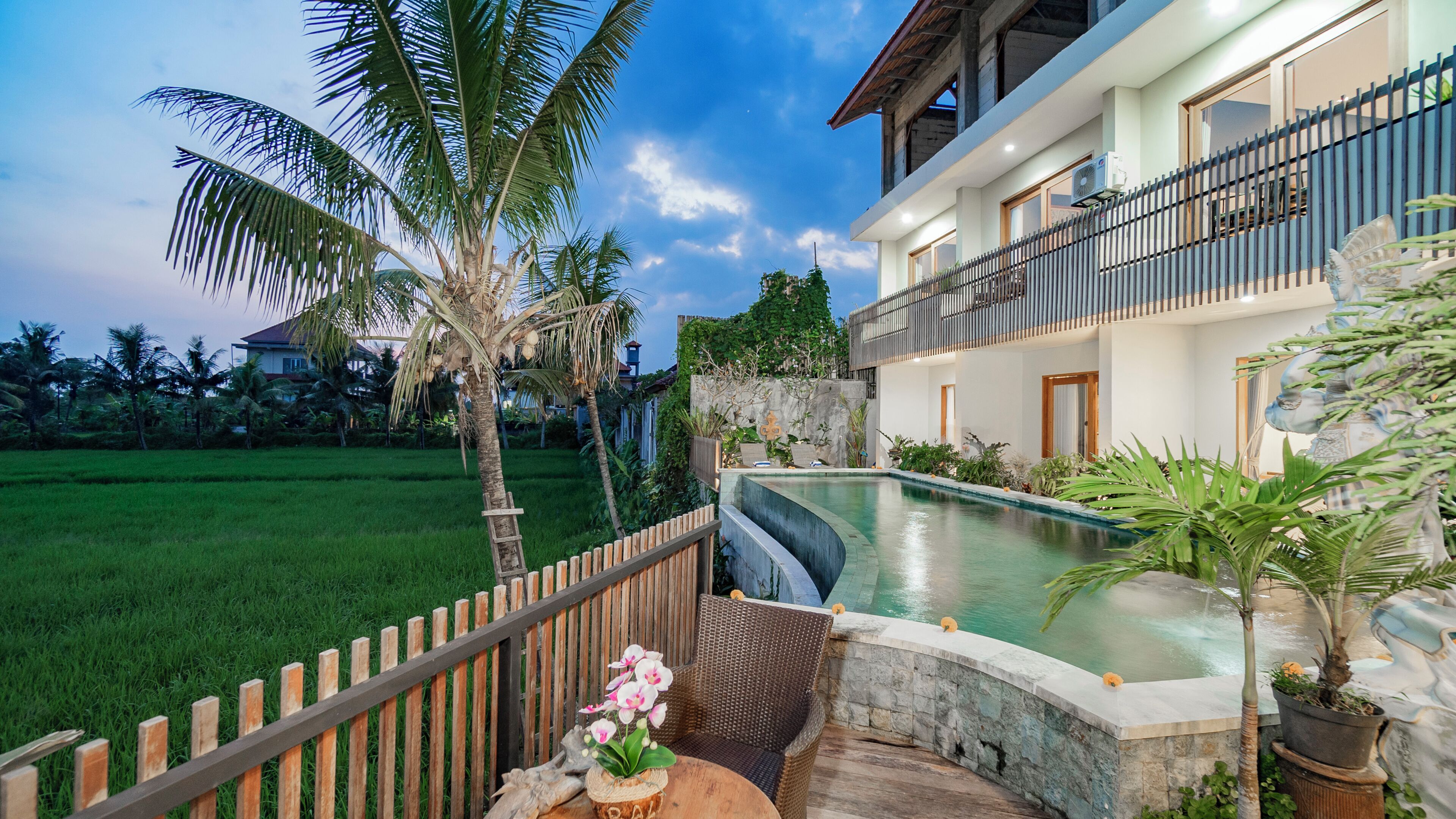 A two-story house with a swimming pool, a wooden fence, and a garden with palm trees.