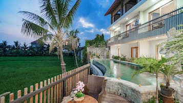 A two-story house with a swimming pool, a wooden fence, and a garden with palm trees.