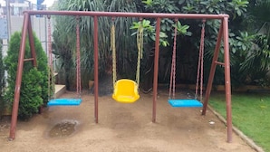 Columpio con dos asientos, uno amarillo y otro azul, en un jardín con diversas plantas.