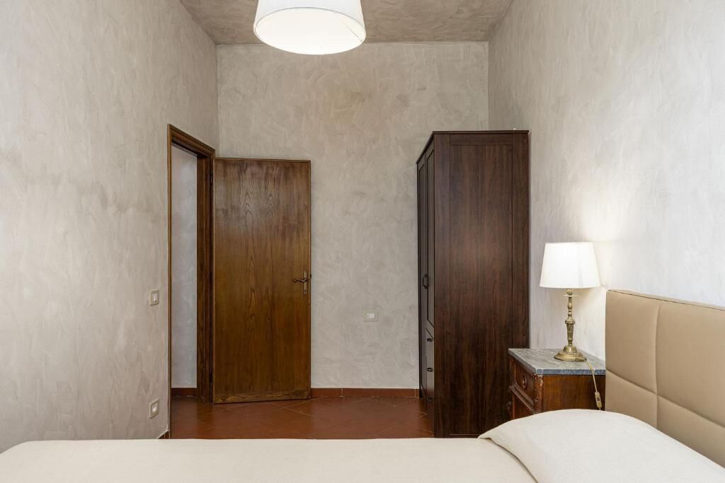 A bedroom with a bed, a nightstand with a lamp, a wardrobe, and a door leading to another room.