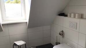 Ein kleines Badezimmer mit Toilette, Mülleimer, Toilettenpapierhalter und einem Fenster mit Blick auf Bäume.
