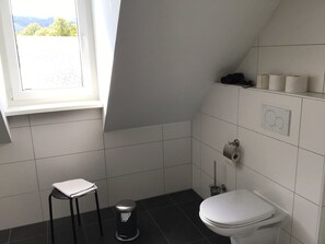 A small bathroom with a toilet, a trash bin, a toilet paper holder, and a window with a view of trees. - Just like home, but in a completely different place (Bengel)