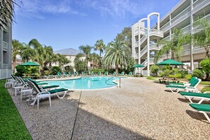 A pool area with lounge chairs and umbrellas, surrounded by palm trees and a multi-story building. - Poolside Vibes, Beach Strolls! Galveston Getaway (Galveston)