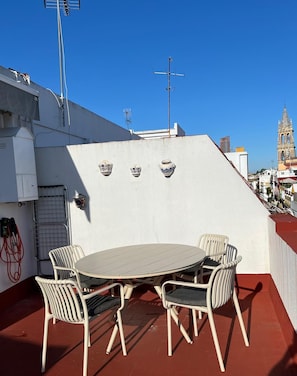 Eine Dachterrasse mit einem runden Tisch und vier Stühlen, eine weiße Wand mit drei Keramiktöpfen und einem strahlend blauen Himmel.