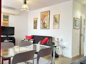 Ein modernes Wohnzimmer mit einer Couch, einem Esstisch mit Stühlen, einem Fernseher und gerahmten Postern an der Wand.