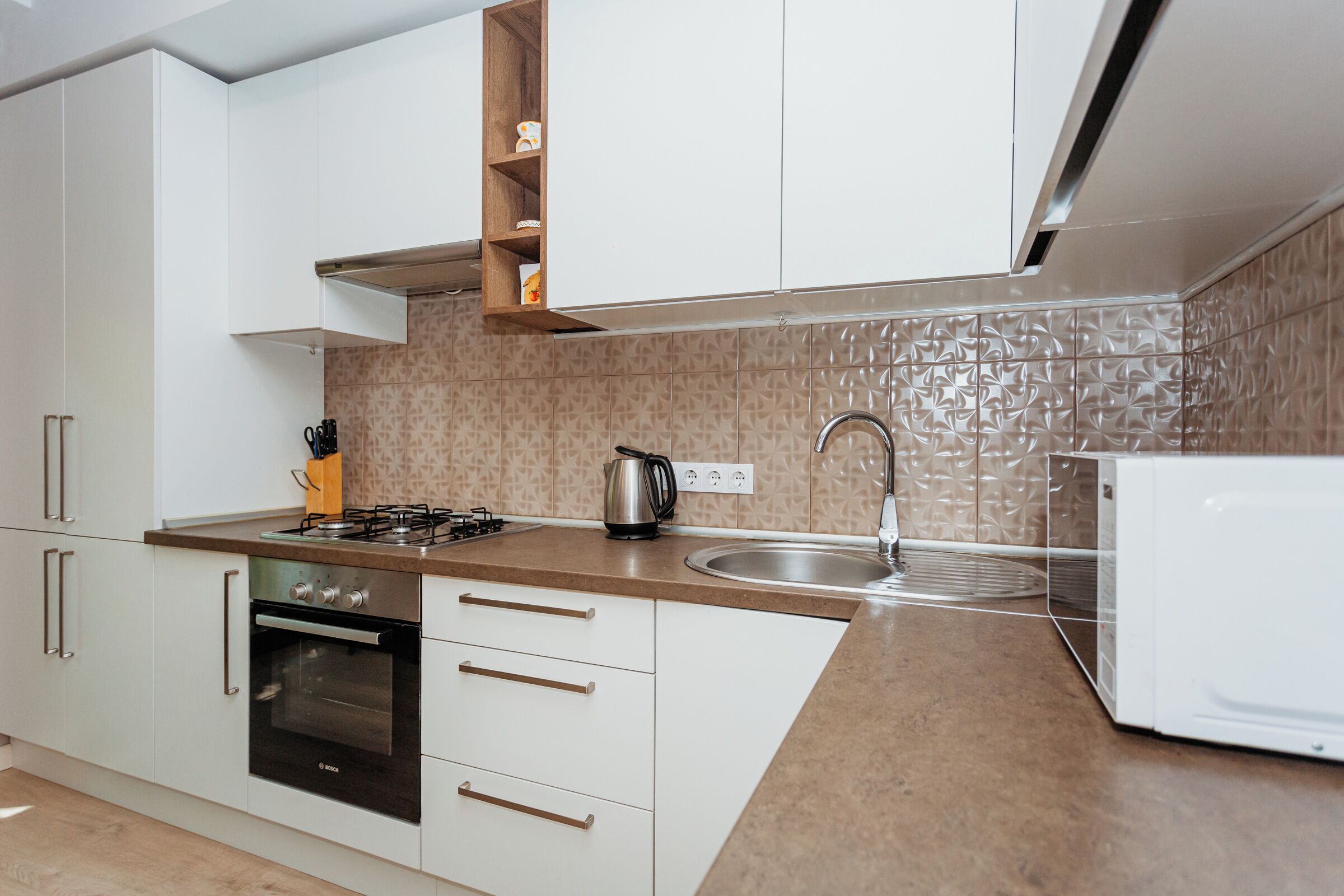Une cuisine moderne avec des meubles blancs, un four Bosch, une cuisinière à gaz, une bouilloire et un évier.