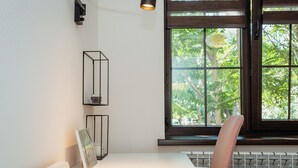 Ein Zimmer mit einem weißen Tisch, einem Stuhl, einem Heizkörper, einem Fenster mit Blick auf Bäume und einer Wandleuchte.