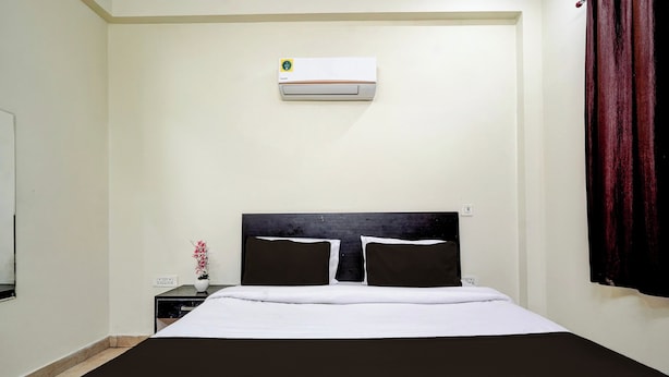 Una habitación de hotel con cama, mesita de noche, un florero con flores, unidad de aire acondicionado y ventana con cortinas.