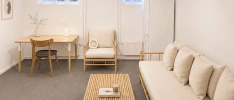 A room with a wooden sofa, a small table, a chair, and a desk with a lamp. There are windows with blinds, a radiator, and a picture on the wall.