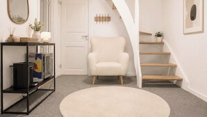 A modern interior with a round beige rug, a white armchair, a black shelf with items, a white door, and a staircase.