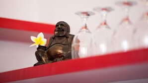 Una estatua con una flor, sobre un fondo rojo con copas de vino desenfocadas.