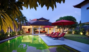 A swimming pool with lounge chairs and umbrellas, surrounded by greenery and a building with a tiled roof. - Villa Alma (Badung)