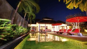 Un área junto a la piscina con sillones de descanso y sombrillas, rodeada de palmeras y un edificio con techo tradicional.