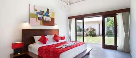 Une chambre d’hôtel avec un grand lit, un mur d’accent rouge, un ventilateur de plafond et une vue sur le jardin, visible à travers la porte-fenêtre coulissante.