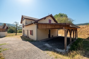 A two-story house with a wooden porch, a gravel driveway, and a rural landscape in the background.