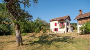 Uma casa de dois andares com telhado vermelho e paredes brancas, rodeada por árvores e grama.