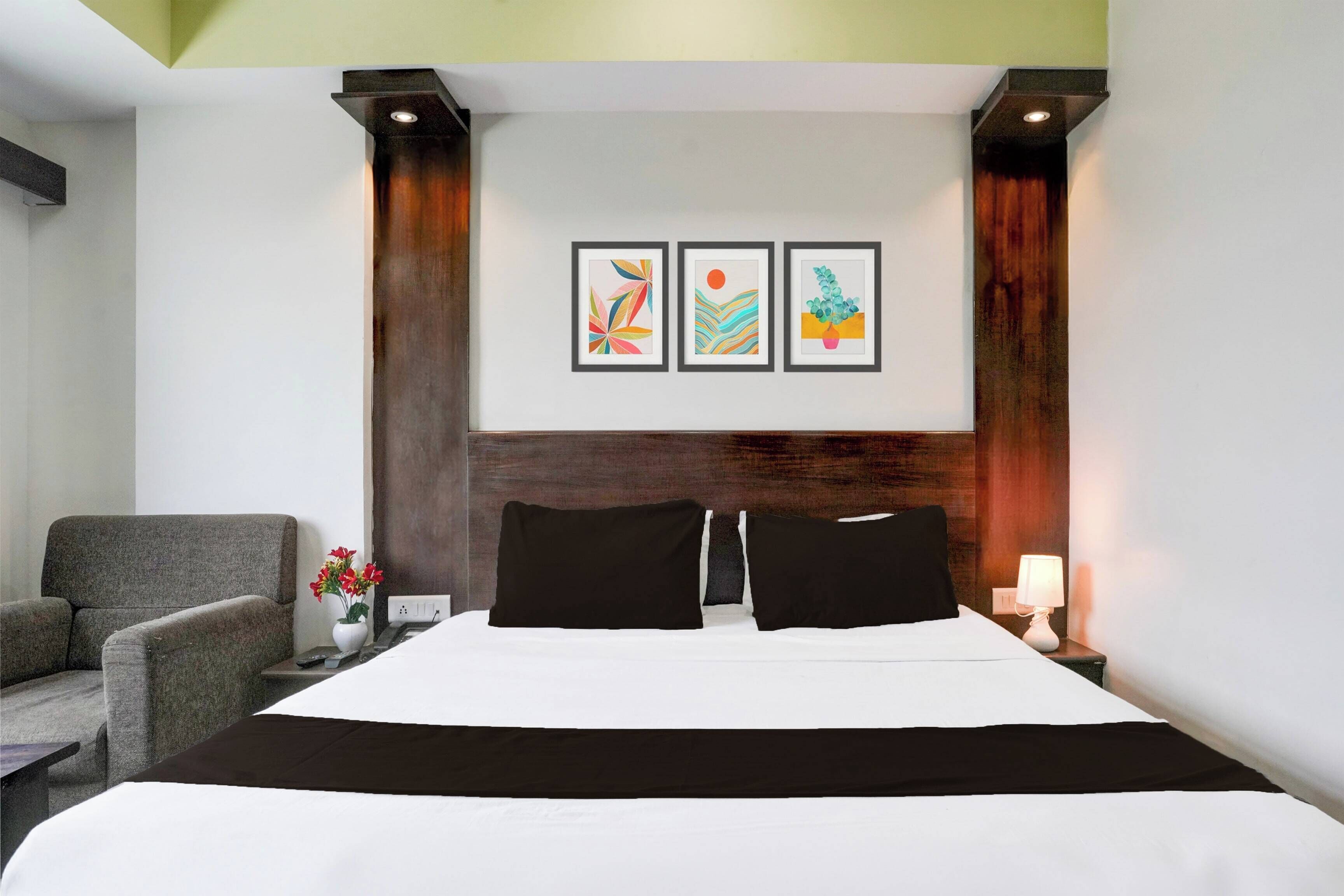 Habitación de hotel con una cama, una silla, una mesita de noche con lámpara y tres obras de arte enmarcadas en la pared.