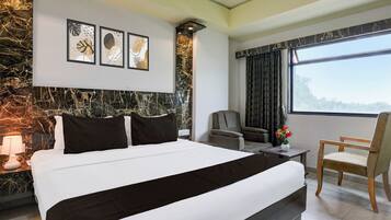 A modern hotel room with a large bed, a seating area with a sofa and chairs, a desk, and a large window offering a view.