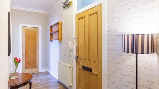 A hallway with a wooden door, a radiator, a wall-mounted phone, and a table with a vase of flowers. - Serene 2BDR Flat Near Edinburgh Old Town (Edinburgh)