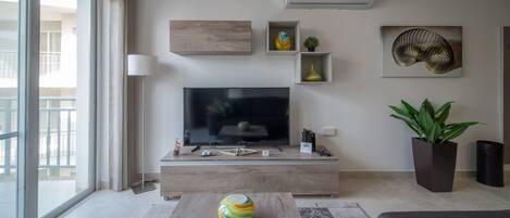 Una sala moderna con una televisión de pantalla plana, una mesa de centro con objetos de colores y una planta grande en maceta.