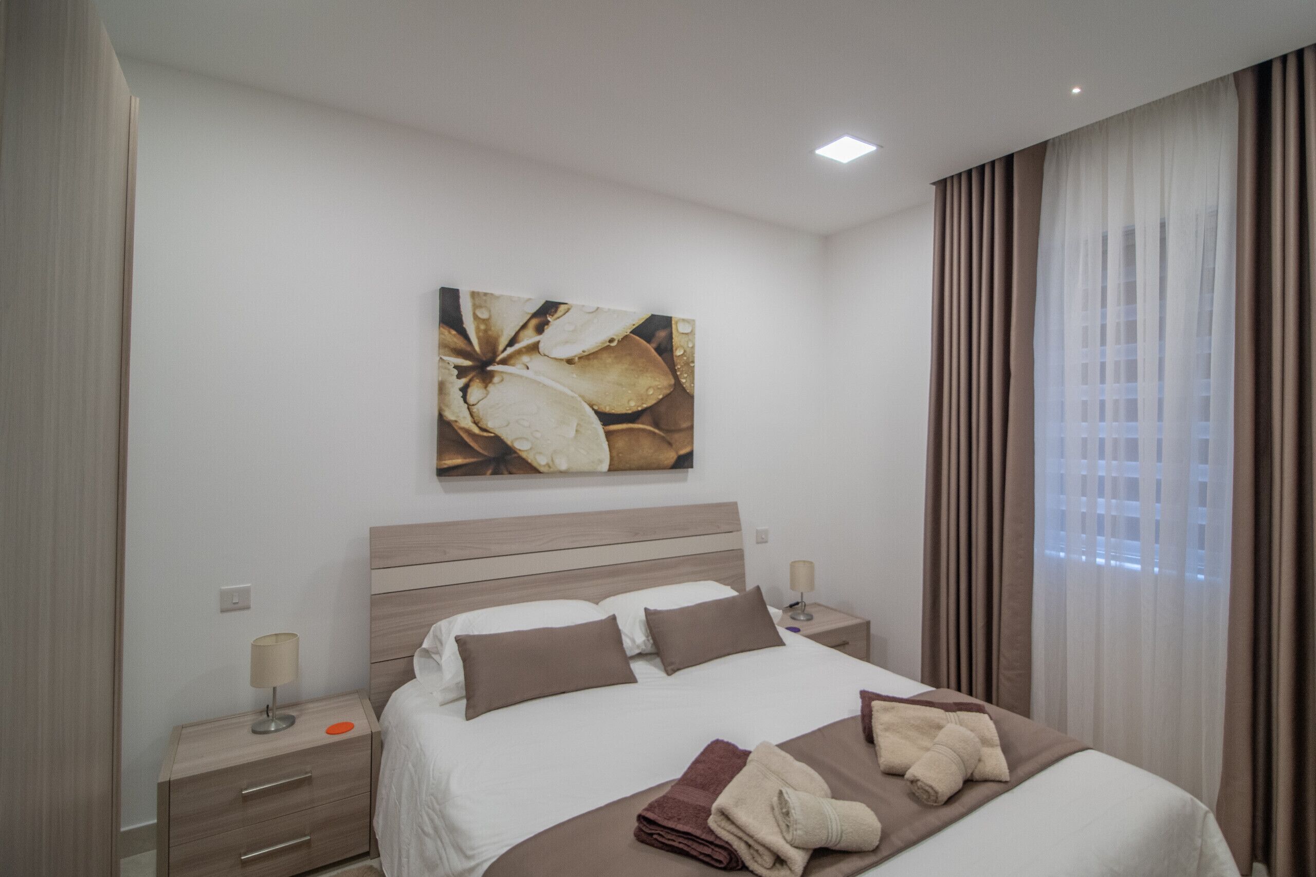 Habitación de hotel con cama, mesita de noche, lámpara, un cuadro y ventana con cortinas.